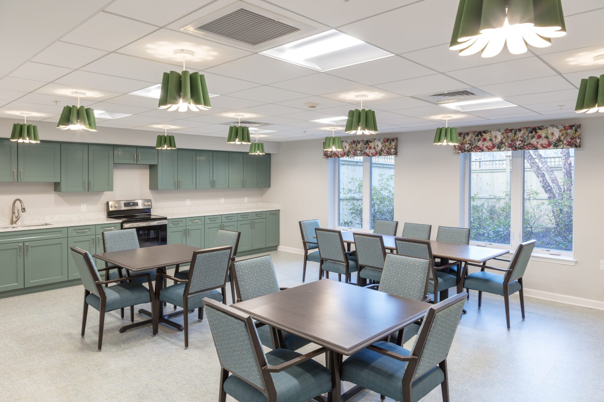 A bright modern kitchen and dining area with teal cabinetry and matching chairs, floral curtains, and pendant lights