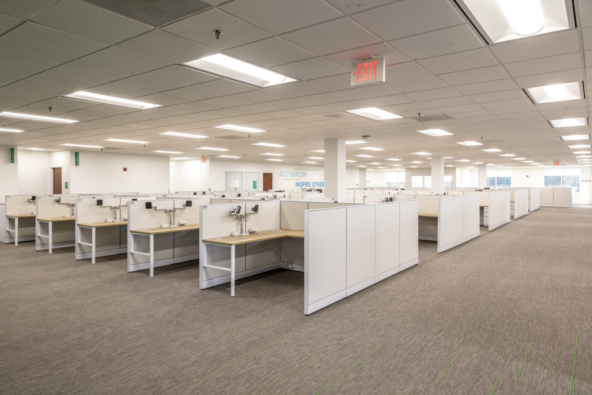 Modern office space with rows of empty cubicles, bright lighting, and an "ACCOMPLISH INSPIRE OTHER" sign