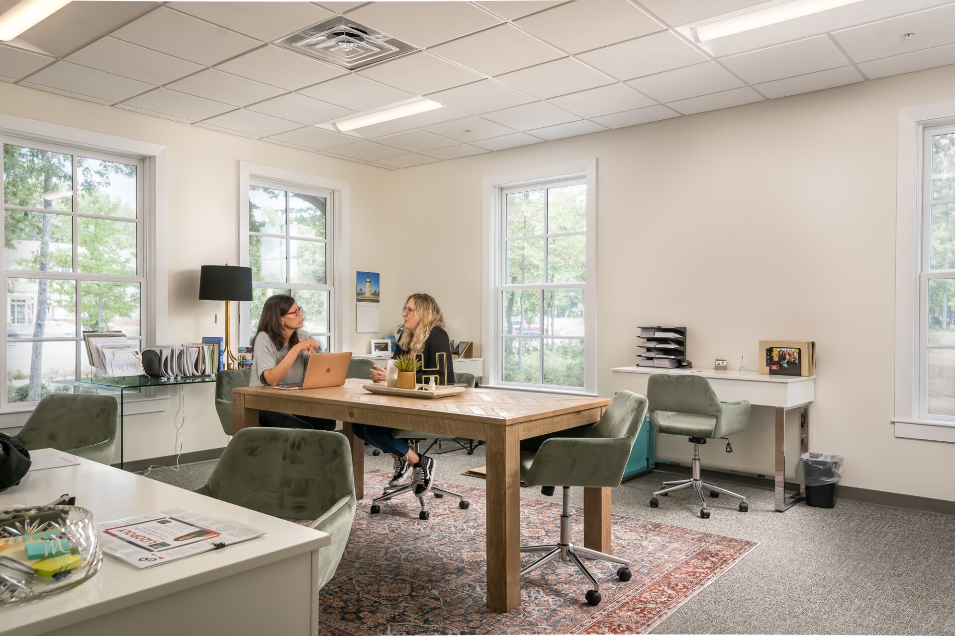 Two people working at a table in a brightly lit office with large windows and modern decor